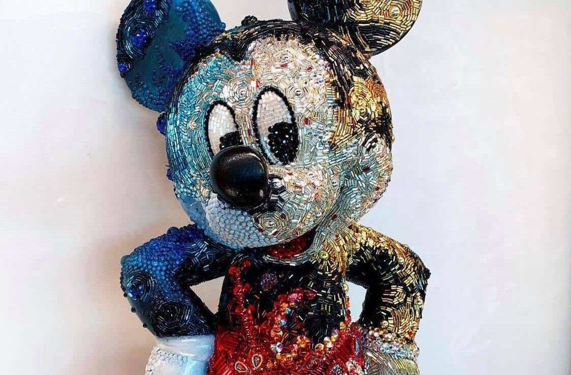 A whimsical beaded Mickey Mouse sculpture featuring his signature blue ear, black nose, and bright red outfit stands out against a plain wall—an eye-catching tribute to Disney magic from Creations Shop.