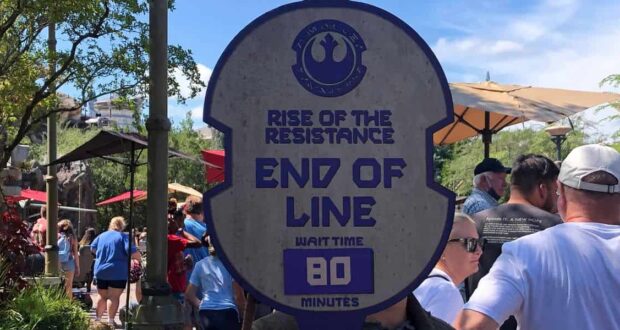 A theme park employee holds a sign reading “Rise of the Resistance End of Line, Wait Time 80 Minutes” in front of a line of people outdoors on a sunny day.