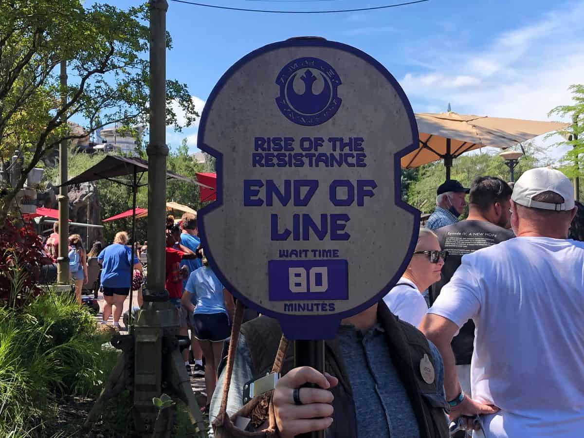 A theme park employee holds a sign reading “Rise of the Resistance End of Line, Wait Time 80 Minutes” in front of a line of people outdoors on a sunny day.