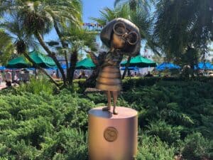 Edna Mode bronze statue, complete with her signature glasses, stands among palm trees and umbrellas in a lush garden at Disney's Hollywood Studios as part of the Fab 50 Character Collection.