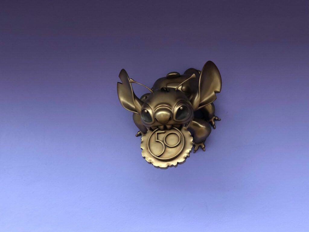 A playful bronze Toy Story alien sculpture clutching a shimmering "50" medallion pops against a purple backdrop, marking the magical celebration of Walt Disney World’s 50th Anniversary.