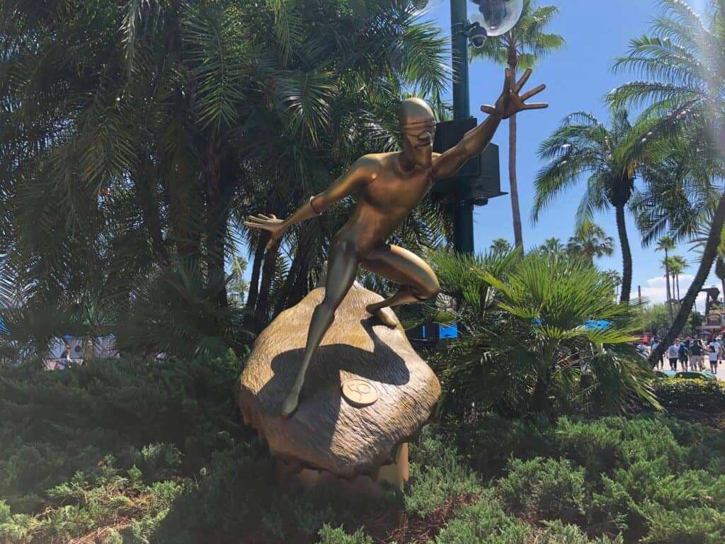 The stunning golden statue of the Silver Surfer glistens in the sunlight amid tropical palm trees and lush greenery, an iconic piece from the Fab 50 Character Collection at Disney’s Hollywood Studios.
