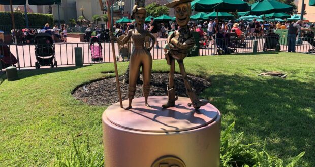 Woody and Jessie from the Fab 50 Character Collection gleam in gold as they pose atop a round pedestal adorned with a "50" emblem, sparkling under the sun at Disney’s Hollywood Studios.