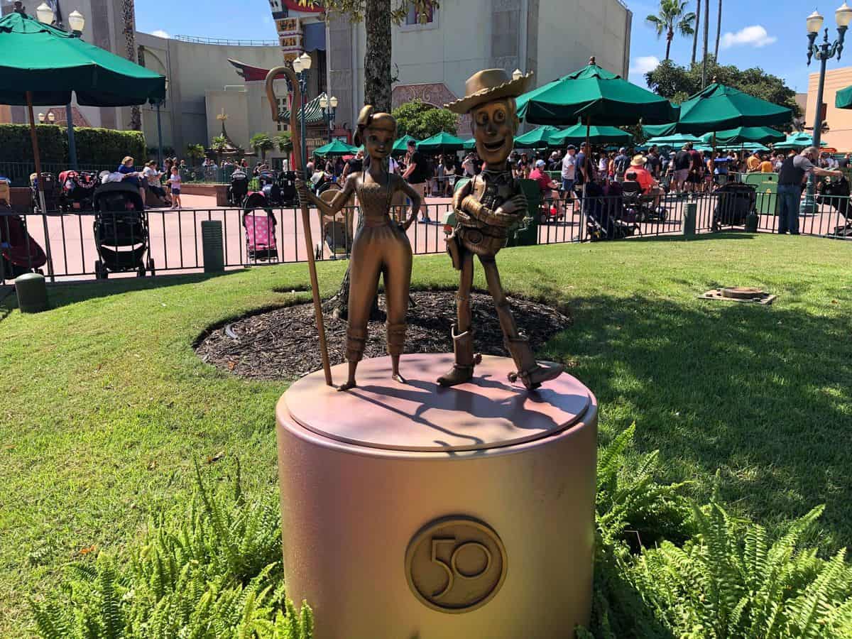 Woody and Jessie from the Fab 50 Character Collection gleam in gold as they pose atop a round pedestal adorned with a "50" emblem, sparkling under the sun at Disney’s Hollywood Studios.