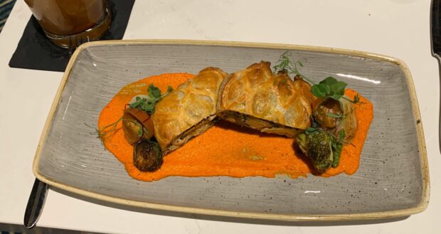 A delicious pastry dish served at Steakhouse 71 in Walt Disney World is artfully plated on a vibrant orange sauce, topped with fresh greens and Brussels sprouts. The picture-perfect presentation, alongside a refreshing beverage, makes it an ideal meal for Disney foodies looking to savor and snap memorable dining moments during their magical visit.