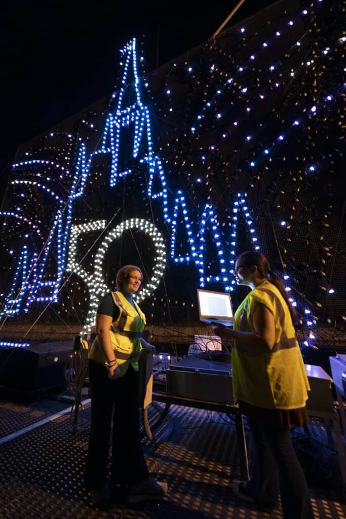 Cast Members in yellow vests work near a glowing "50" and Cinderella Castle display at night, highlighting new scenes unveiled for the 50th Anniversary of Disney’s Electrical Water Pageant.