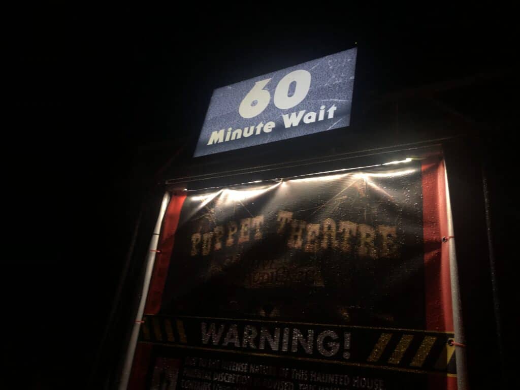 A "60 Minute Wait" sign is displayed above a haunted house facade at Universal’s Halloween Horror Nights, illuminated at night with a bold "Warning!" posted underneath.