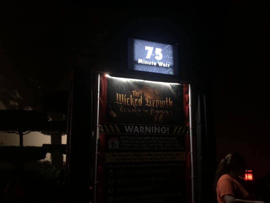 The Wicked Growth: Realm of the Pumpkin" attraction sign at Halloween Horror Nights shows a 75-minute wait time amid nighttime festivities. Guests are reminded to follow all safety procedures while waiting in line.