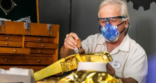 A talented Cast Member, sporting a galaxy-themed mask and protective goggles, carefully paints a golden prop at a workshop lined with wooden drawers, demonstrating the exceptional craftsmanship that brings Disney magic to life.