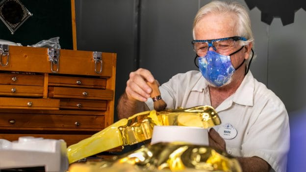 A talented Cast Member, sporting a galaxy-themed mask and protective goggles, carefully paints a golden prop at a workshop lined with wooden drawers, demonstrating the exceptional craftsmanship that brings Disney magic to life.