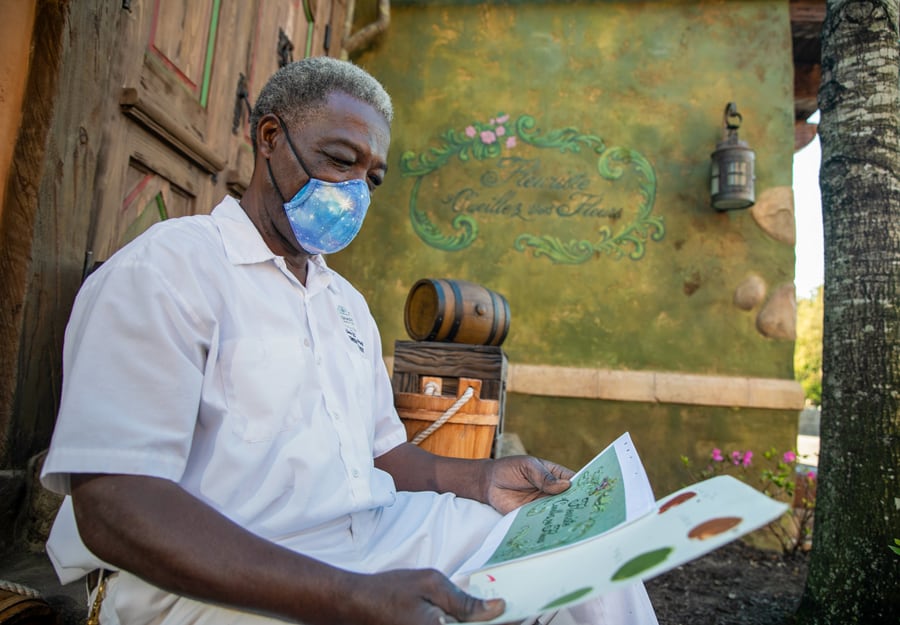 A guest in crisp white attire and a blue face mask reviews the menu outside the rustic, signage-adorned building at Disney’s themed land, showcasing the Imagineers’ meticulous attention to immersive detail.