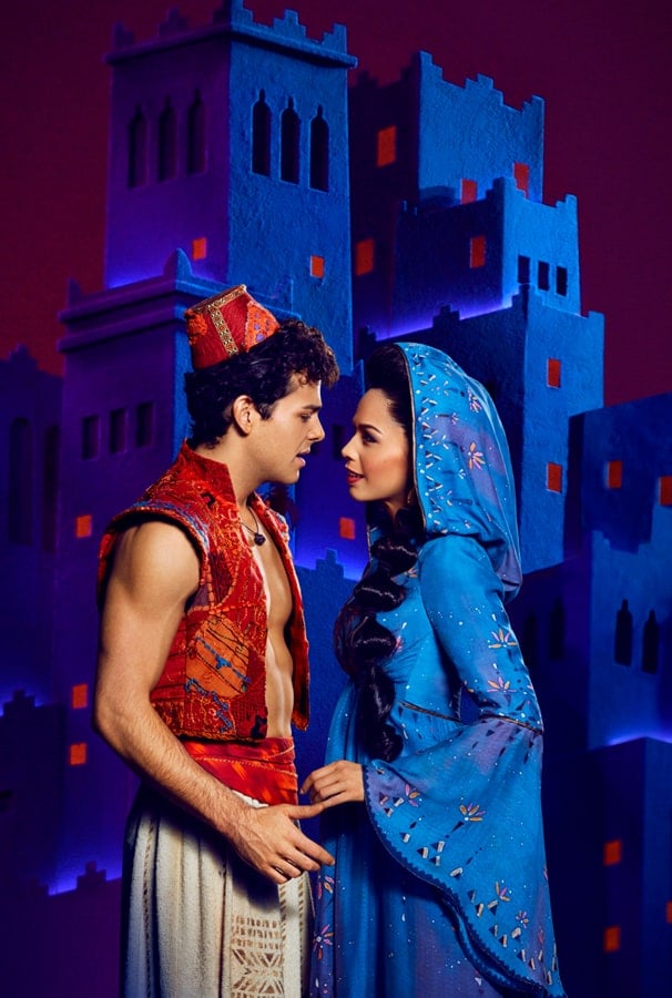 Broadway performers in vibrant costumes share a magical moment in front of a dazzling Disney-inspired blue and purple palace set, capturing the excitement and spectacle of live entertainment at a Disney park.