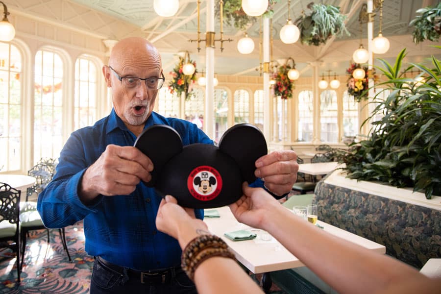 Mickey Mouse surprises a guest with iconic ears inside a dazzling, plant-filled Disney World restaurant adorned with chandeliers, as the magic of Disney and the Class of 1971 are joyfully celebrated.