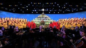 During the EPCOT Candlelight Processional, choir members in vibrant red and gold robes create a stunning human Christmas tree on stage, with the orchestra performing festive melodies in front under shimmering blue lights—a dazzling holiday tradition at Walt Disney World captured on November 26.