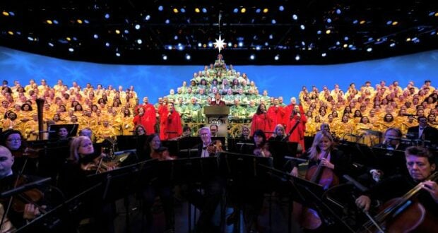 During the EPCOT Candlelight Processional, choir members in vibrant red and gold robes create a stunning human Christmas tree on stage, with the orchestra performing festive melodies in front under shimmering blue lights—a dazzling holiday tradition at Walt Disney World captured on November 26.