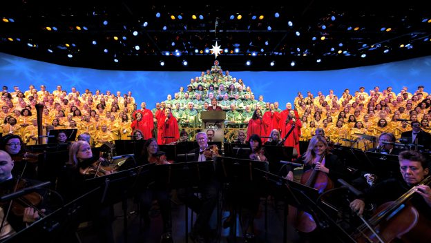 During the EPCOT Candlelight Processional, choir members in vibrant red and gold robes create a stunning human Christmas tree on stage, with the orchestra performing festive melodies in front under shimmering blue lights—a dazzling holiday tradition at Walt Disney World captured on November 26.
