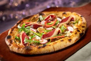 California Grill-inspired artisan pizza served on a rustic wooden board, topped with fresh greens, radish slices, red onions, and thin cuts of cured meat—an eye-catching treat crafted just for Walt Disney World’s 50th Anniversary celebration.