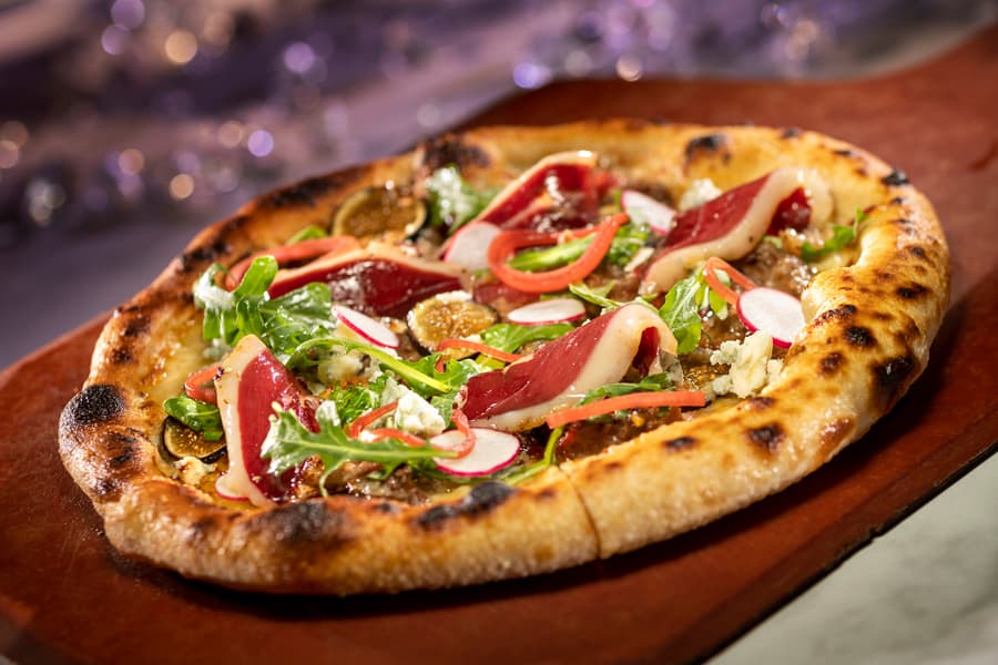 California Grill-inspired artisan pizza served on a rustic wooden board, topped with fresh greens, radish slices, red onions, and thin cuts of cured meat—an eye-catching treat crafted just for Walt Disney World’s 50th Anniversary celebration.