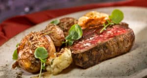 A perfectly cooked medium-rare steak served with roasted potatoes, cauliflower, and fresh greens on a speckled plate—this signature entrée pays homage to California Grill's gourmet flair, specially crafted for Walt Disney World's magical 50th Anniversary celebration.
