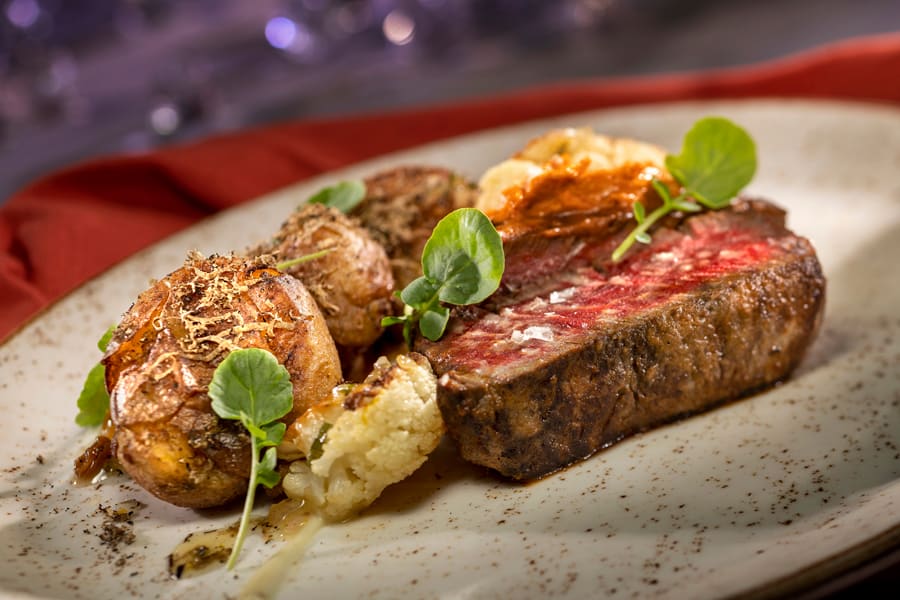 A perfectly cooked medium-rare steak served with roasted potatoes, cauliflower, and fresh greens on a speckled plate—this signature entrée pays homage to California Grill's gourmet flair, specially crafted for Walt Disney World's magical 50th Anniversary celebration.