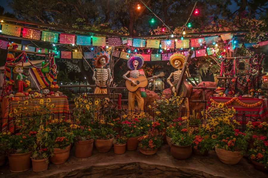 Día de los Muertos comes alive at Disneyland Resort with festive skeleton figures performing music surrounded by vibrant flowers and dazzling colorful lights, creating an immersive nighttime celebration for guests.