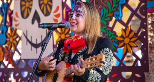 A Día de los Muertos performer with intricate face paint sings and plays guitar on stage, surrounded by lively, colorful decorations that capture the festive energy of Disneyland Resort's seasonal celebrations.