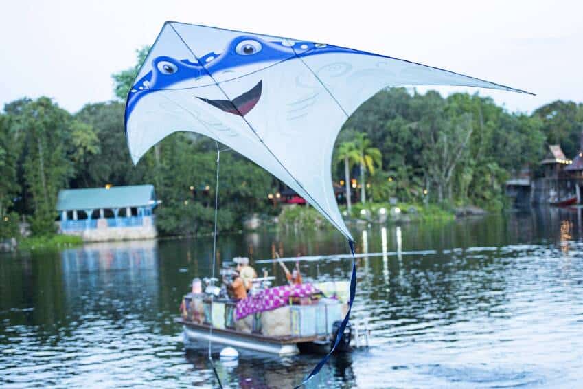 The KiteTales blue manta ray kite, beaming with a friendly smile, soars above a boat cruising on a serene lake framed by lush trees—inviting guests to experience the enchanting outdoor magic of the park's adventure zone.