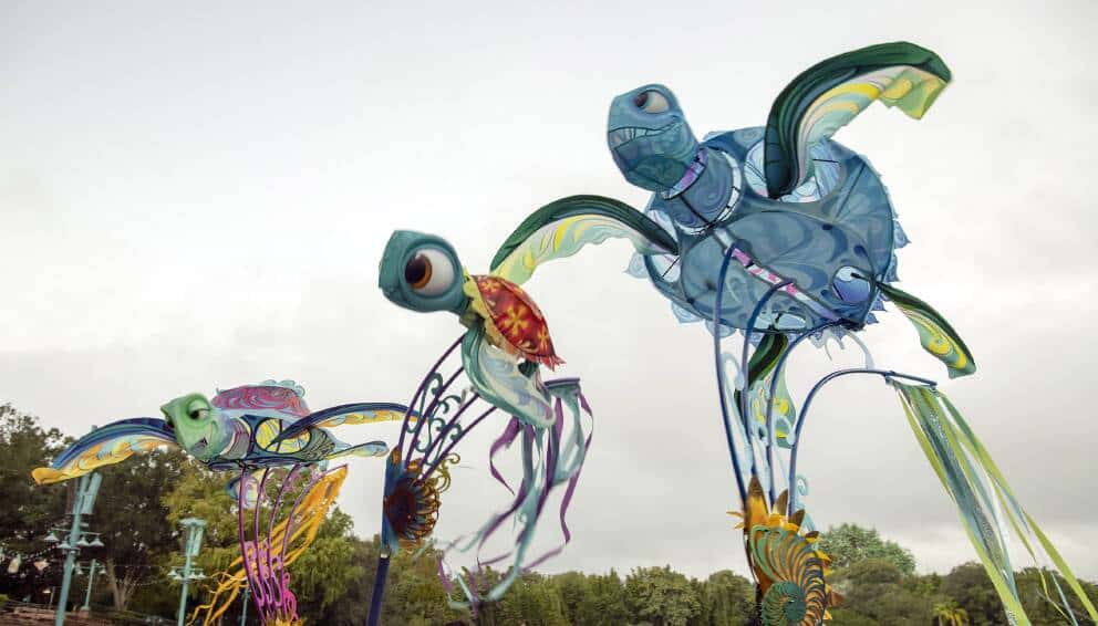 Three vibrant sea turtle puppets from KiteTales soar above the audience at Disney’s Animal Kingdom, set against lush trees and a dramatic cloudy sky, creating an enchanting moment in this imaginative park spectacle.