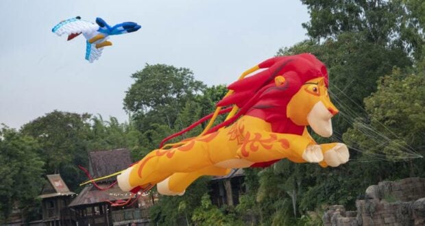 Marvel at the magic of KiteTales as giant lion and blue bird kites gracefully glide above the treetops and buildings along Discovery River at Disney’s Animal Kingdom, filling the sky with vibrant color and wonder.