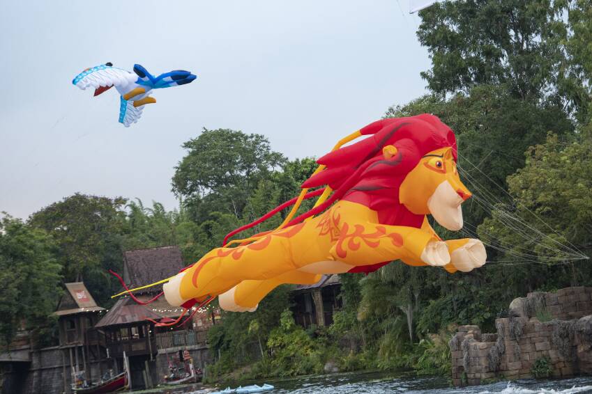Marvel at the magic of KiteTales as giant lion and blue bird kites gracefully glide above the treetops and buildings along Discovery River at Disney’s Animal Kingdom, filling the sky with vibrant color and wonder.