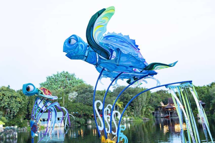 Two vibrant blue sea turtle sculptures soar above the Discovery River at Disney’s Animal Kingdom, framed by lush trees and park buildings, showcasing the dynamic artistry of KiteTales.