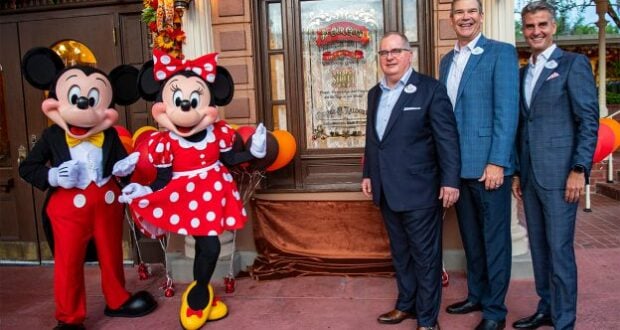 Mickey Mouse and Minnie Mouse, joined by three Disney Cast Members in suits, pose beside a special commemorative plaque outside a festively decorated building at Walt Disney World, surrounded by vibrant fall decorations that highlight the joyful atmosphere of the celebration.