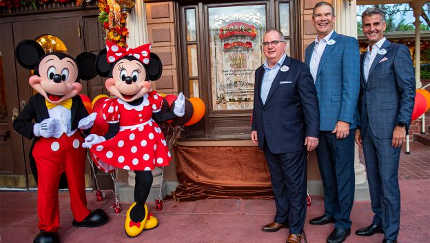 Mickey Mouse and Minnie Mouse, joined by three Disney Cast Members in suits, pose beside a special commemorative plaque outside a festively decorated building at Walt Disney World, surrounded by vibrant fall decorations that highlight the joyful atmosphere of the celebration.