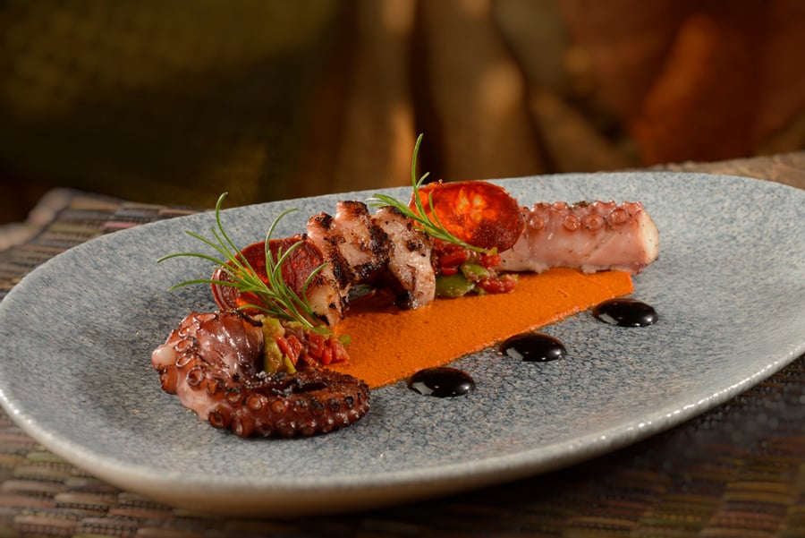 Grilled octopus with savory sauce, topped with herbs and slices of chorizo, is masterfully arranged on a vibrant blue plate, echoing the festive spirit and vivid hues of the Coco scene from Mickey's PhilharMagic attraction.