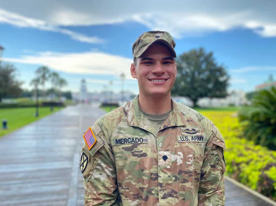 A proud U.S. Army soldier, beaming in uniform, pauses for a photo along the boardwalk at a military appreciation day event, lush greenery and dramatic clouds setting the perfect backdrop.