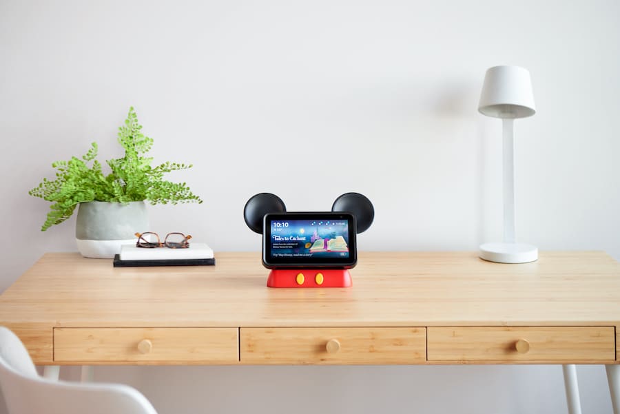 A Mickey Mouse-inspired smart display brightens up a wooden desk alongside a plant, books, and a white lamp—bringing the magic of Disney Parks into your home with every glance.