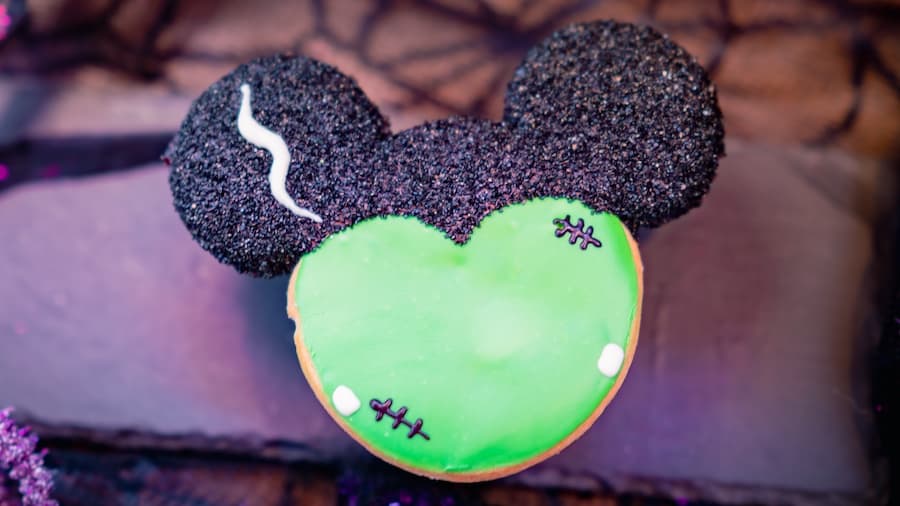 Frankenstein Mickey Donut, topped with green icing and black sugar Mickey ears, rests on a dark surface—an irresistible Disneyland treat perfect for fans of Disney parks and spooky delicious snacks.