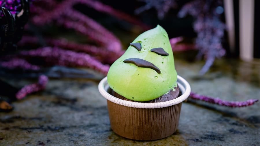 A Disneyland chocolate cupcake with playful green frosting shaped into a smiling face, topped with black candy eyes and mouth—a whimsical treat that brings a touch of Disney magic and delightful flavor to fans exploring the park’s enchanting offerings.