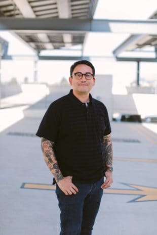 A Disney cast member with glasses and tattoos stands on a rooftop in a black shirt and jeans, facing the camera and reflecting the creative spirit that energizes Disney Parks entertainment teams worldwide.