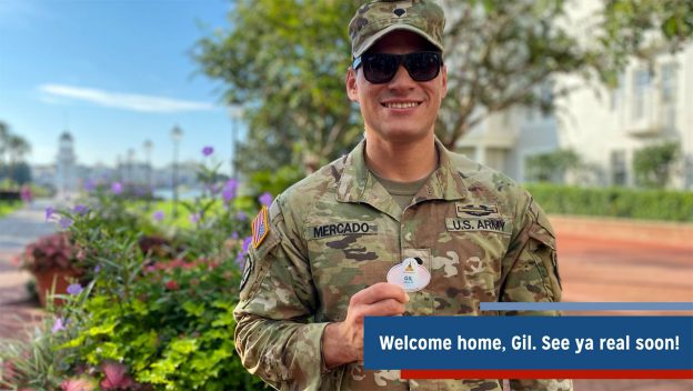 A U.S. Army soldier, all smiles in full uniform, stands outdoors at Disney Parks holding a celebratory Disney pin and a “Welcome home” message—capturing the heartfelt magic of a hero’s return to the Happiest Place on Earth.