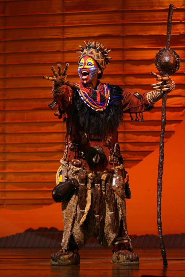 A Broadway actor in a dazzling tribal-inspired costume and striking face paint commands the stage with a ceremonial staff, set against a vivid orange backdrop that radiates the dynamic excitement of Disney’s celebrated theater productions.