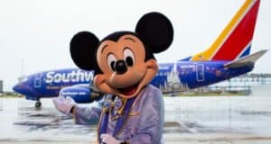 Mickey Mouse, dressed in a light blue suit, greets fans in front of a Southwest airplane adorned with dazzling Disney-themed artwork, bringing the wonder of Disney Parks to the sky for an unforgettable Flying Disney Style experience.
