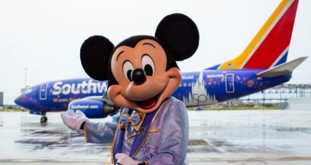 Mickey Mouse, dressed in a light blue suit, greets fans in front of a Southwest airplane adorned with dazzling Disney-themed artwork, bringing the wonder of Disney Parks to the sky for an unforgettable Flying Disney Style experience.