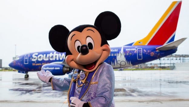 Mickey Mouse, dressed in a light blue suit, greets fans in front of a Southwest airplane adorned with dazzling Disney-themed artwork, bringing the wonder of Disney Parks to the sky for an unforgettable Flying Disney Style experience.