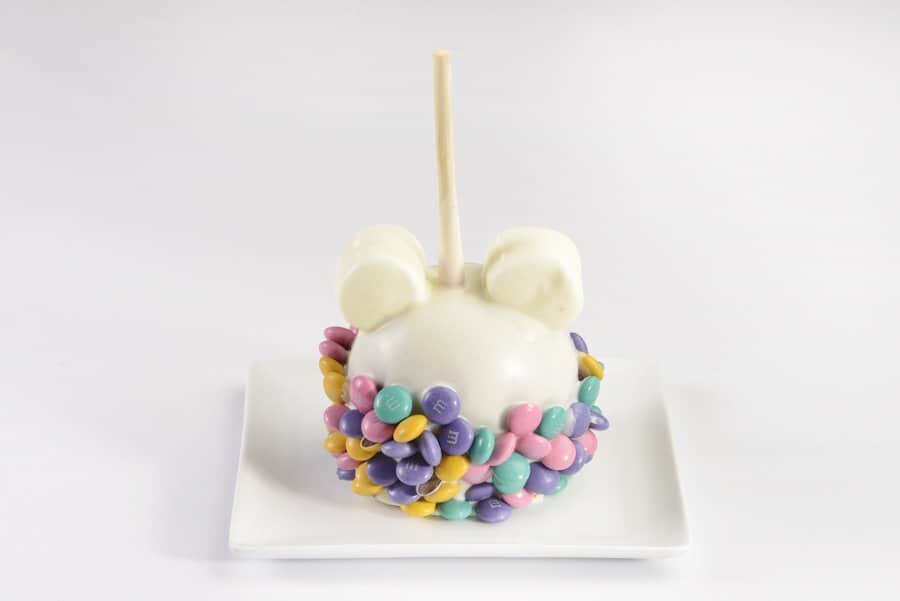 A playful white candy apple topped with candy ears and vibrant sprinkles sits on a white plate, capturing the magical spirit of Main Street Confectionery at Magic Kingdom Park—an irresistible Disney treat sure to delight theme park fans.