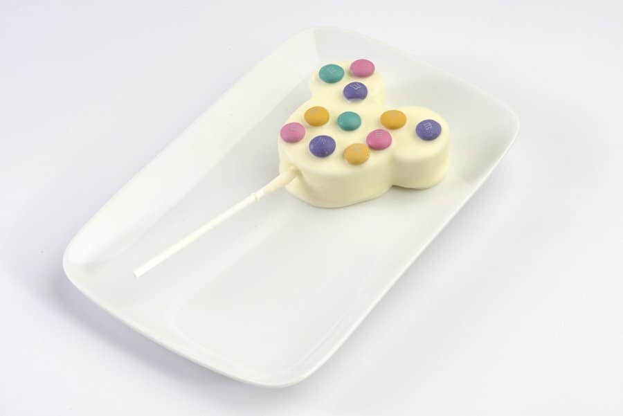 A whimsical white chocolate pop topped with vibrant candy dots, fresh from Main Street Confectionery—now open again at Magic Kingdom Park—served on a crisp white plate, ready for Disney fans to enjoy.