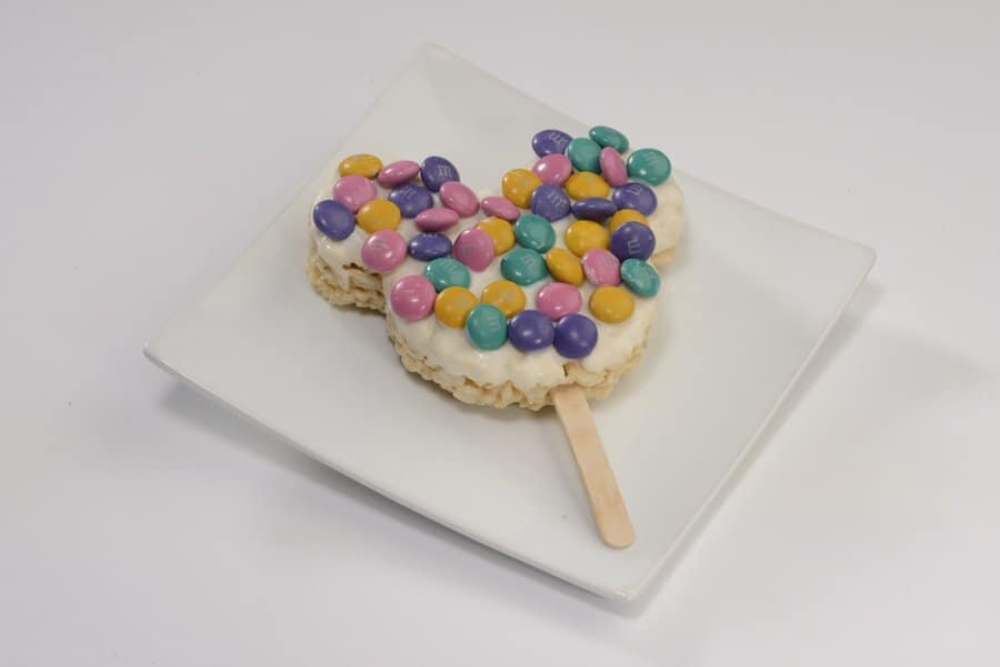 A festive Mickey Mouse-shaped Rice Krispies treat on a stick, covered with colorful candy and served on a white plate—an iconic Main Street Confectionery snack for Disney fans at Magic Kingdom, perfect for celebrating the park’s reopening.