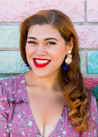 A vibrant individual with long red hair, striking red lipstick, and a floral dress stands joyfully before a lively brick wall backdrop, exuding the same infectious positivity and welcoming spirit that Disney cast members share with guests throughout the parks.