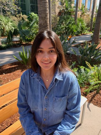 A guest in a denim shirt relaxes on a bench at the heart of a lively theme park, surrounded by lush greenery and palm trees, taking in the energetic atmosphere and colorful entertainment buzzing all around.