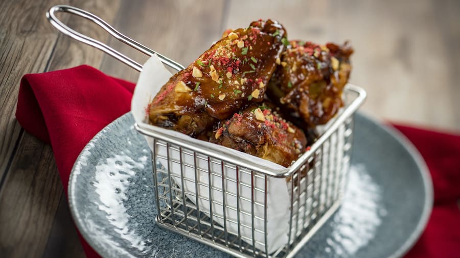 Crispy chicken wings with savory sauce and fresh herbs served in a metal basket on a blue plate with a red napkin, ready to share at Steakhouse 71 inside Disney’s Contemporary Resort—reserve your table to enjoy this must-try treat during your Walt Disney World visit.